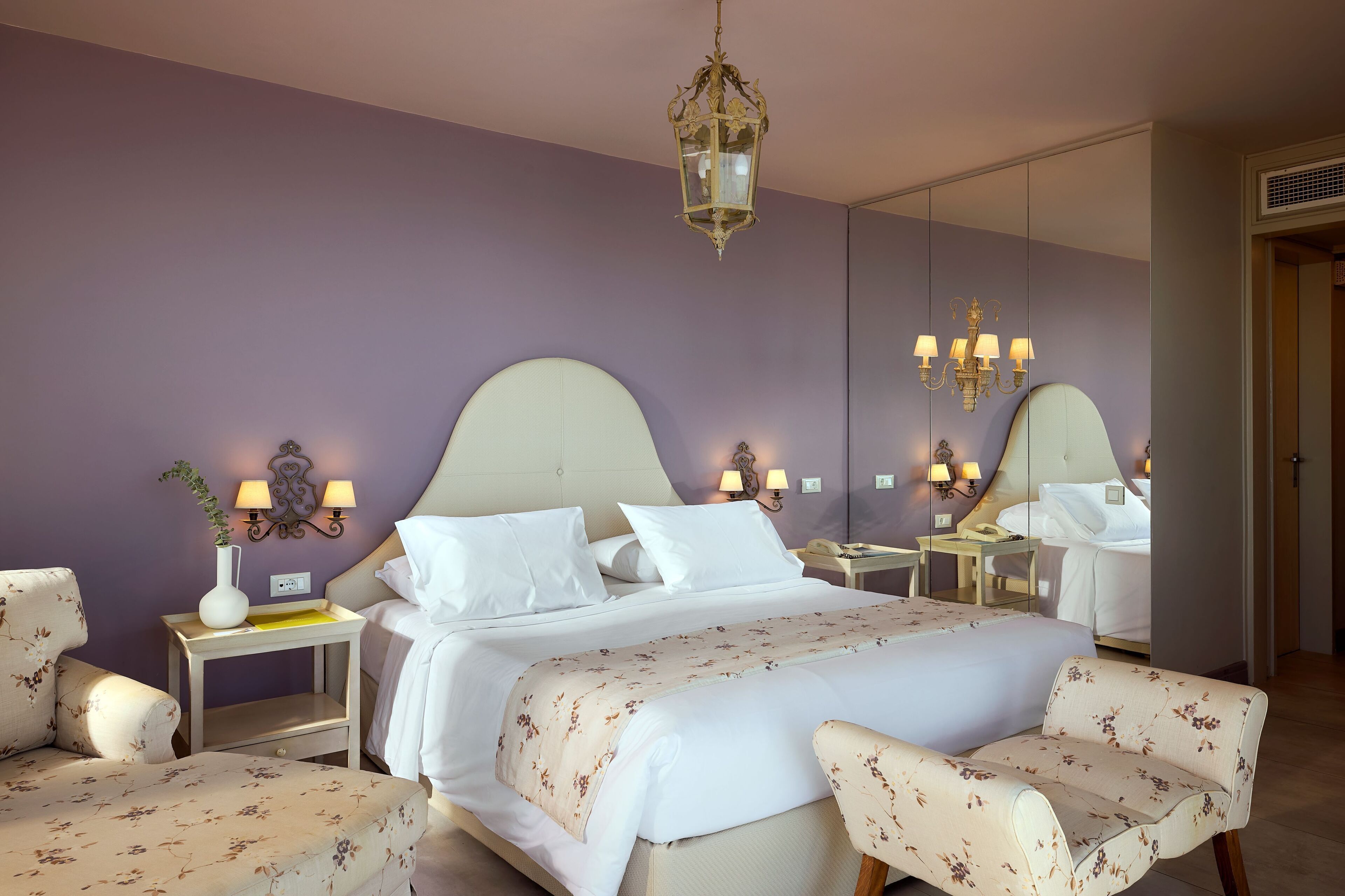 Quarto de hotel com uma cama grande, duas mesas de cabeceira com abajures, um espelho e uma poltrona com padrão floral.