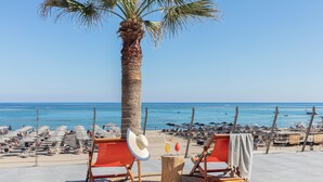 A beachside seating area with a palm tree, two drinks on a wooden table, and beach umbrellas in the background. - Aquila Porto Rethymno (Rethymno)