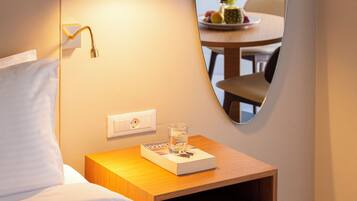 Ein ordentlich bezogenes Bett mit weißen Leinen, ein Nachttisch mit Lampe, ein Spiegel, der einen Essbereich reflektiert, und ein kleiner Tisch mit einem Glas Wasser und einem Buch.