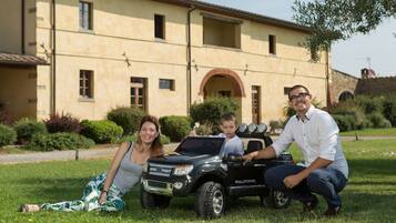 Una família amb un camió de joguina davant d'un edifici gran de dues plantes amb teulada enrajolada i finestres d'arc.