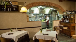 A restaurant interior with tables set for dining, a kitchen area, and a wine display. - Hotel Le Capanne (Arezzo)