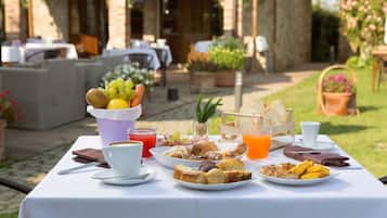 Une table à manger installée à l’extérieur, où sont présentés divers aliments pour le petit-déjeuner, notamment des viennoiseries, des fruits et des boissons.