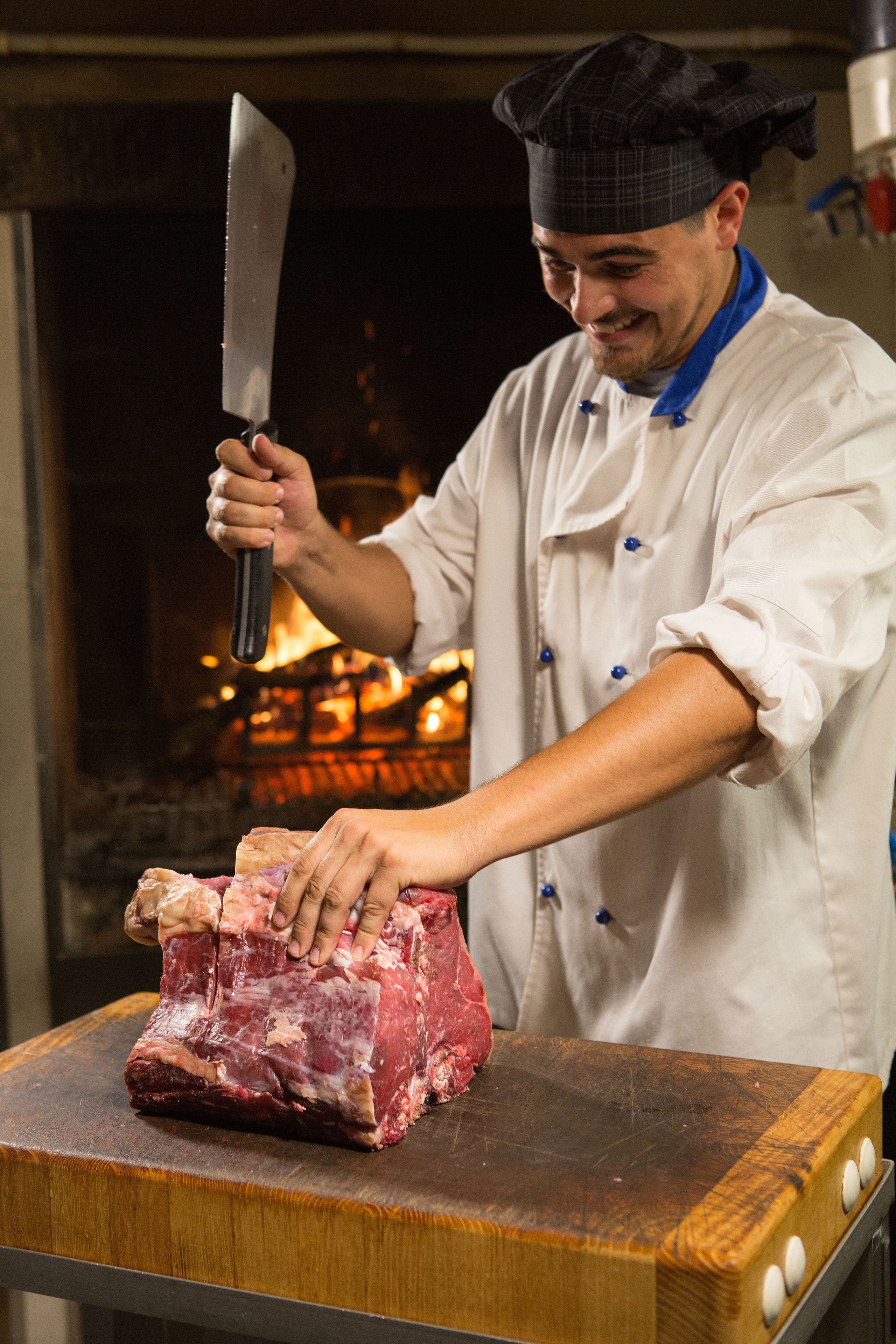 Un chef con uniforme blanco y gorro de cocinero negro sostiene un cuchillo grande y un trozo de carne cruda sobre una tabla de cortar de madera.