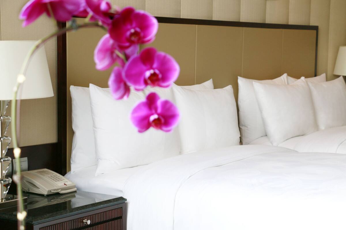 Een hotelkamer met een net opgemaakt bed, een bedtelefoon en een vaas met roze bloemen.