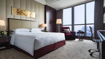 Habitación de hotel con una cama grande, dos mesitas de noche con lámparas, un escritorio con una silla, un sillón rojo y un ventanal con vistas a la ciudad.