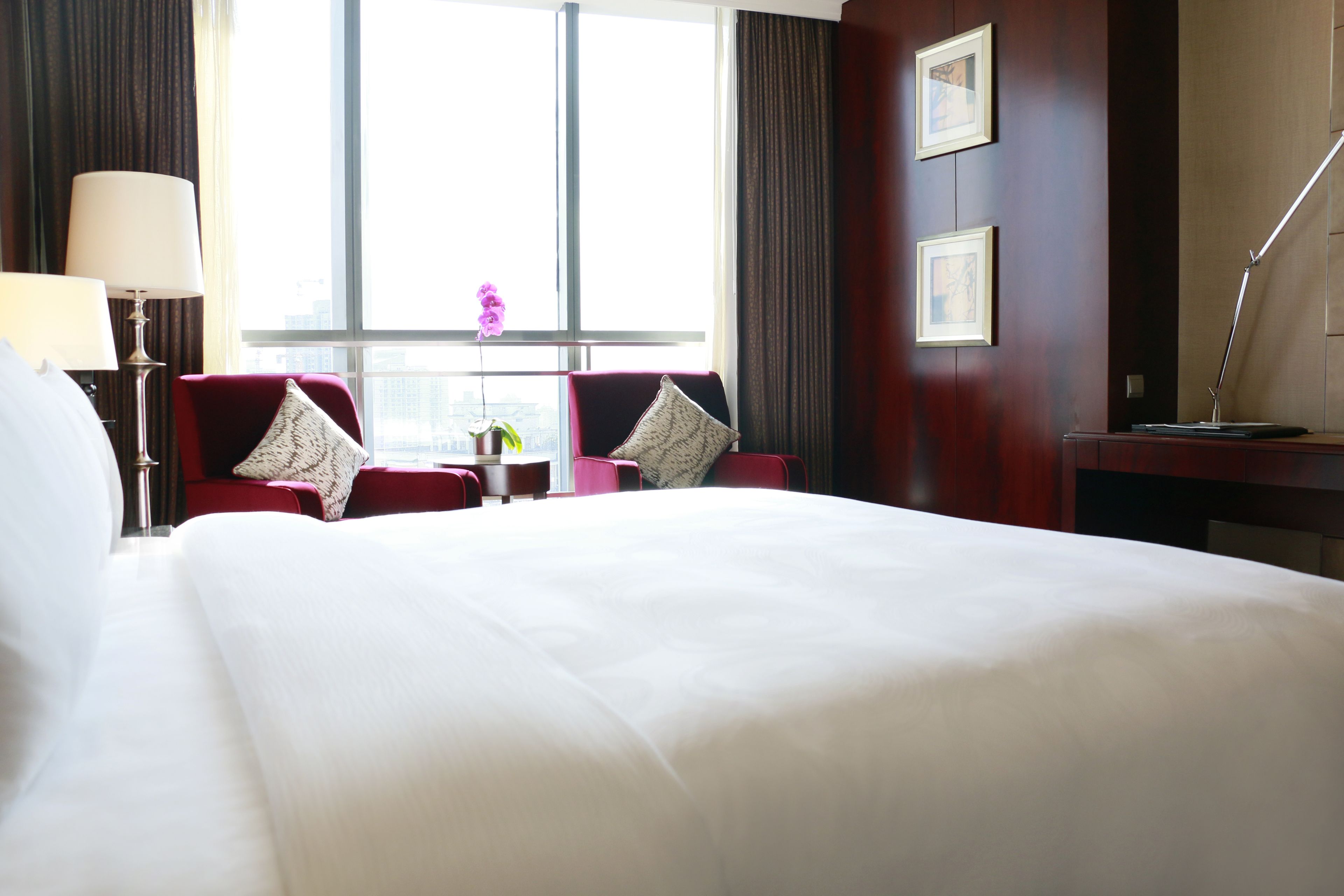 Une chambre d’hôtel avec un grand lit, deux fauteuils rouges, une petite table avec un vase et un bureau avec une lampe.