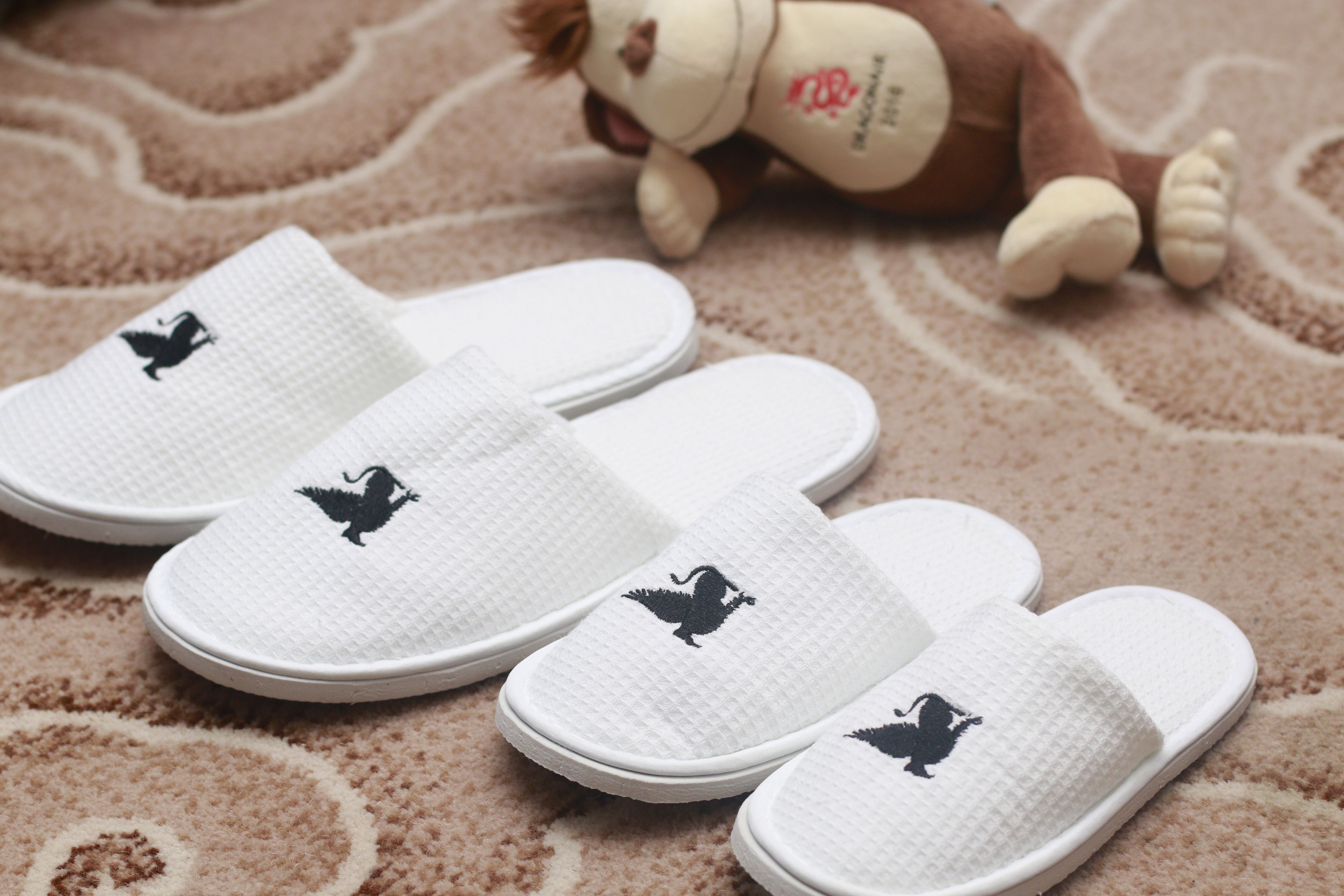 A pair of white slippers with a black bird logo, placed on a patterned carpet.