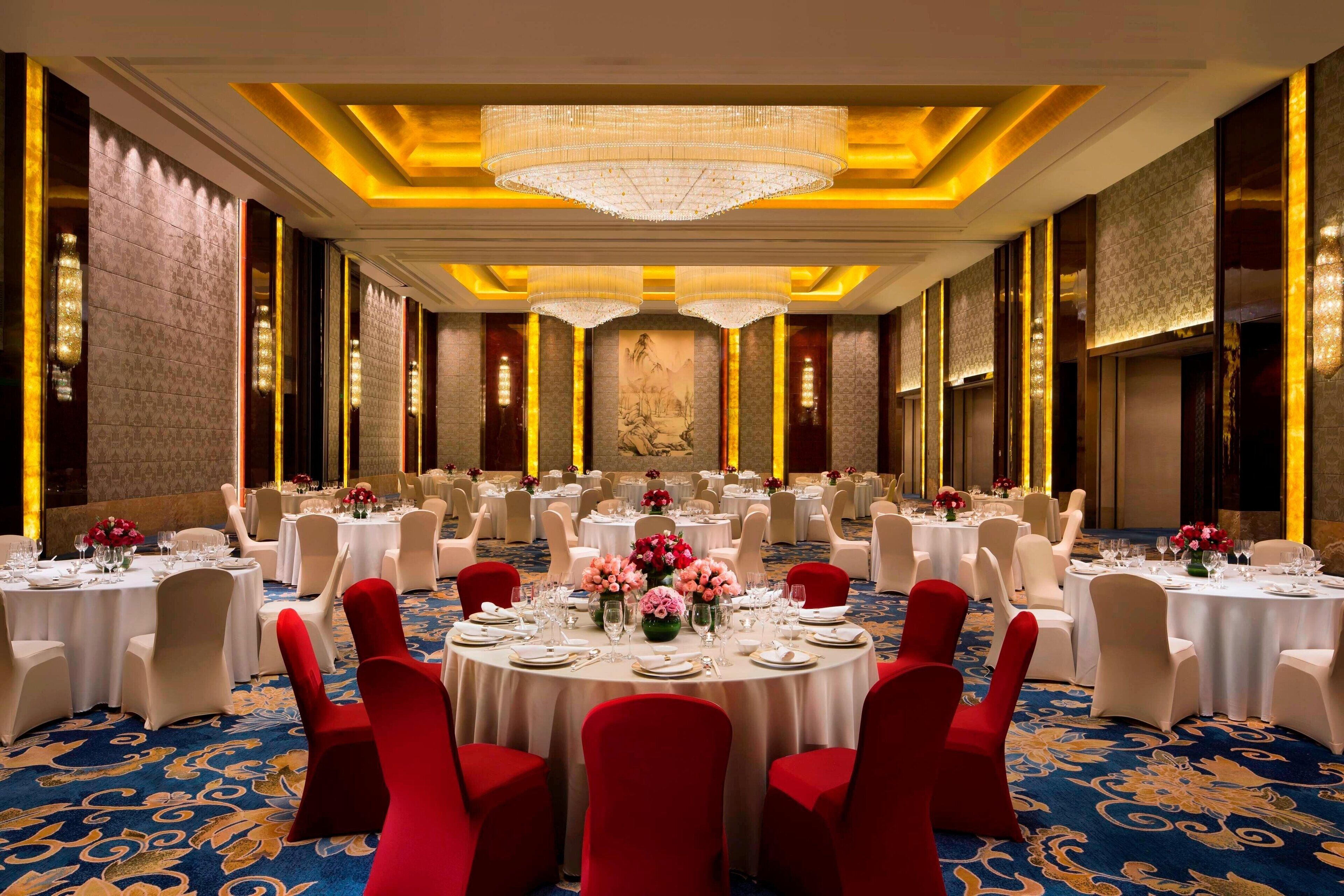 A banquet hall with round tables, red chairs, and chandeliers.