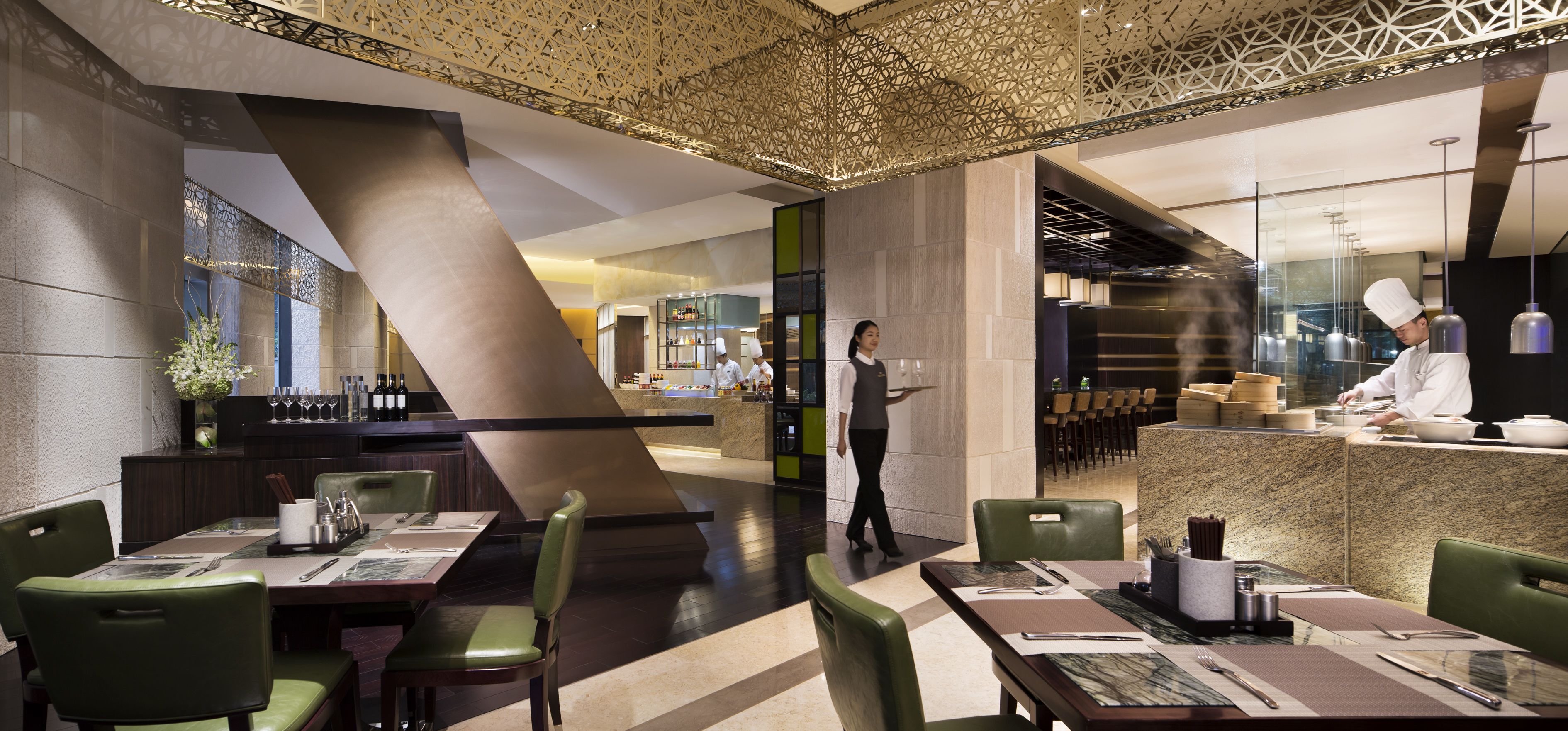 A modern restaurant with a central bar, green upholstered chairs, and a chef preparing food.