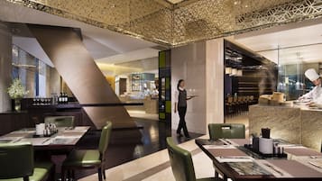 A modern restaurant with a central bar, green upholstered chairs, and a chef preparing food.