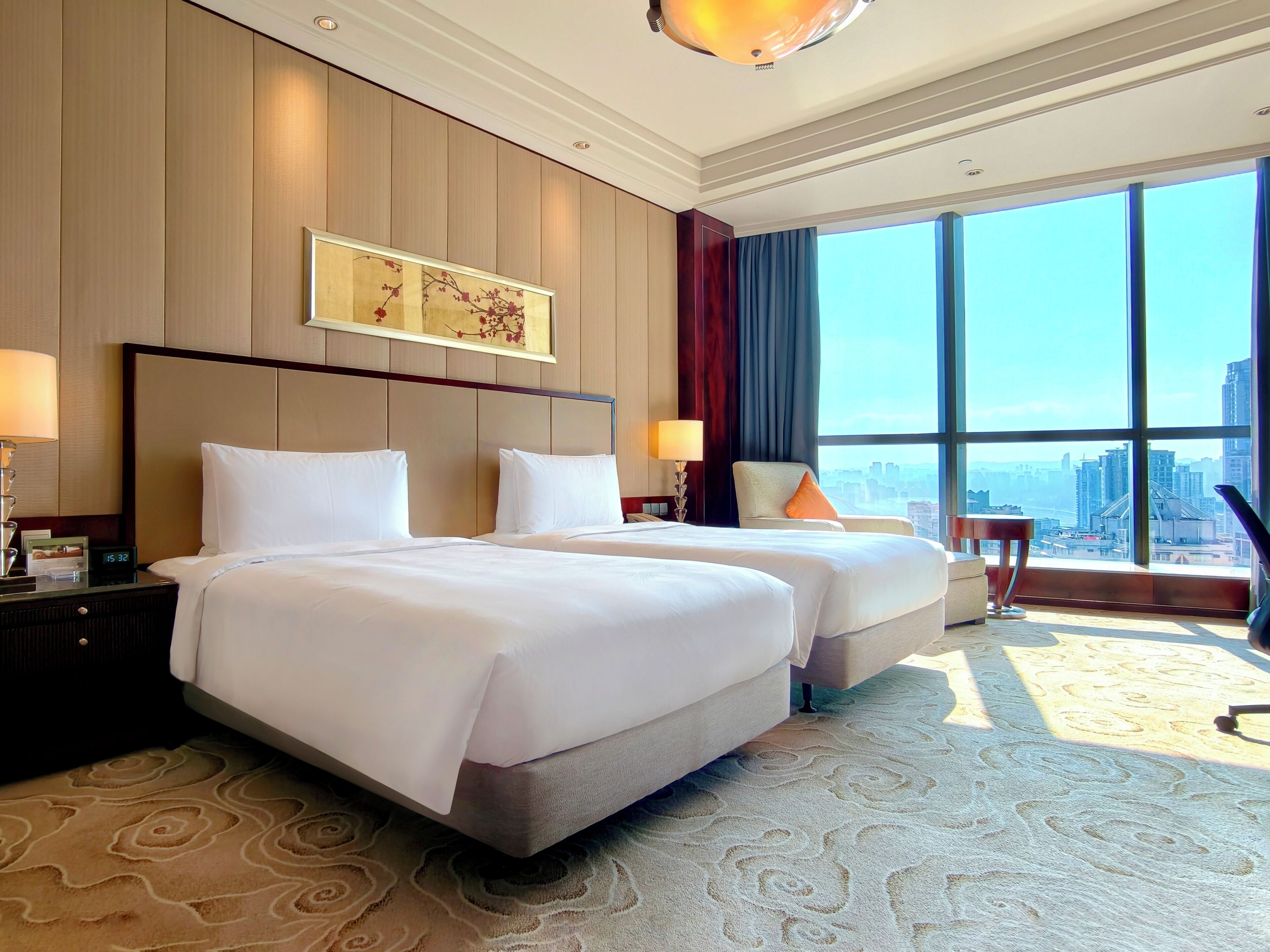 A hotel room with two beds, a large window with a city view, a desk with a lamp, and a chair.