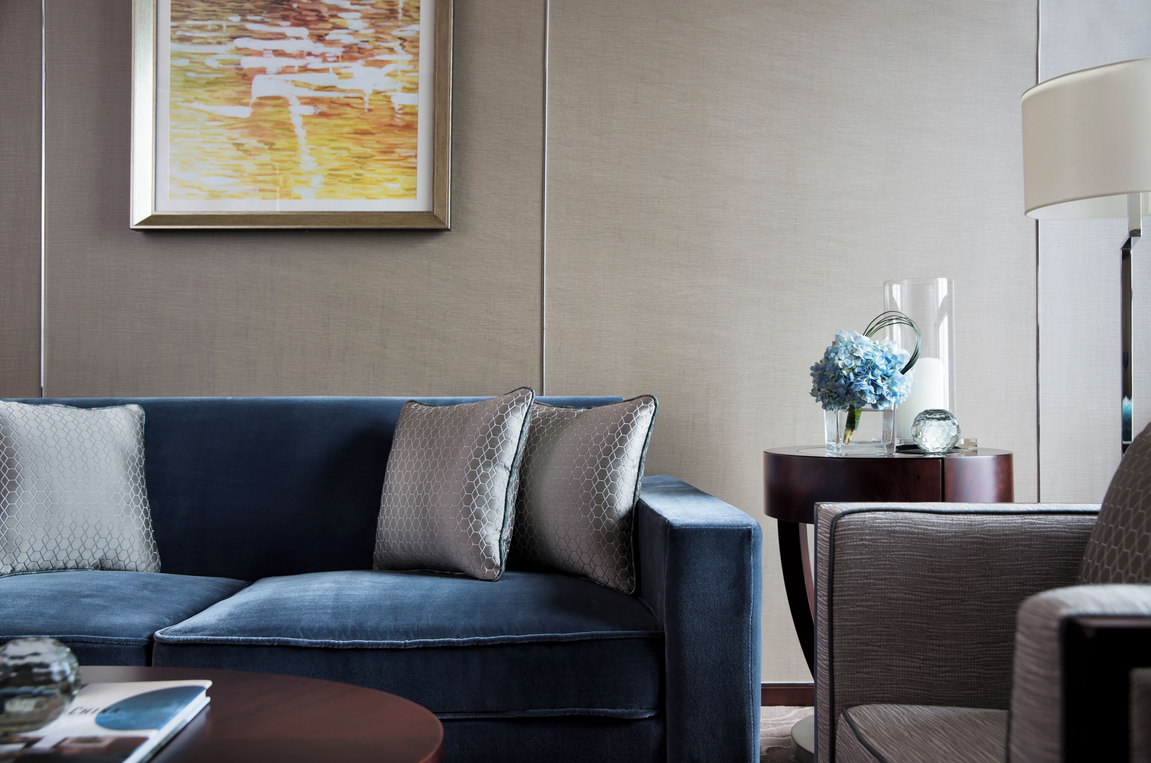 A modern living room with a blue sofa, two decorative pillows, a side table with a vase of flowers, and a lamp.