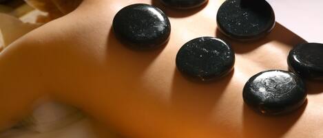A person receiving a hot stone massage with various colored oils and candles on a tray.