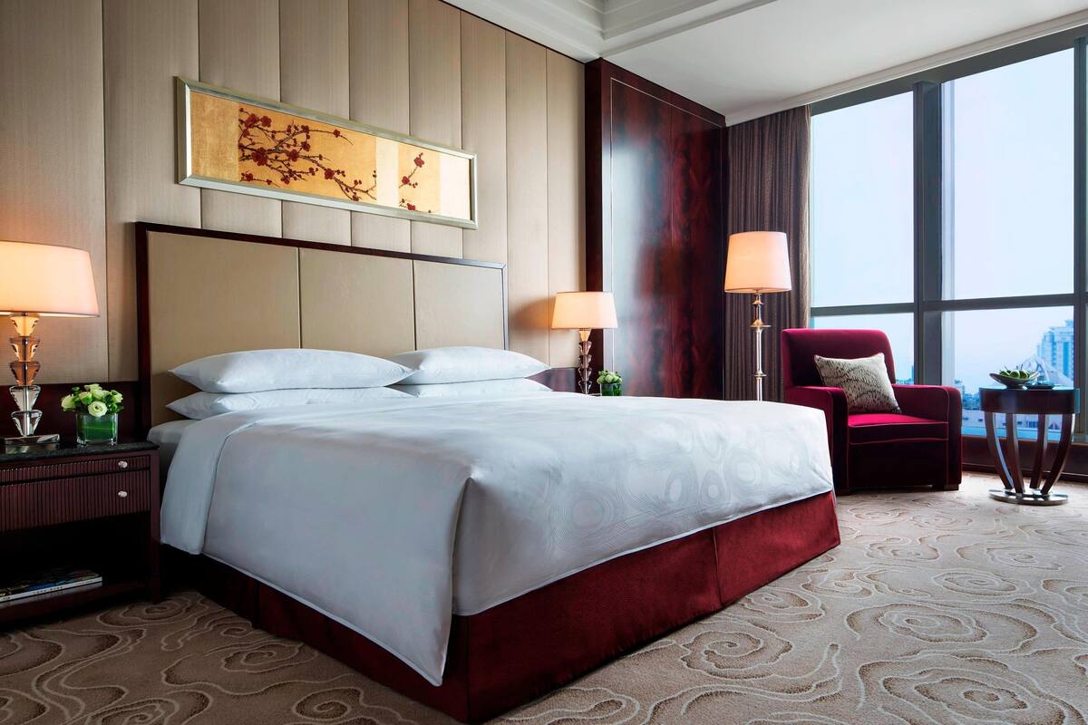 Ein Hotelzimmer mit einem großen Bett, zwei Nachttischlampen, einem roten Sessel und Blick auf die Stadt durch große Fenster.