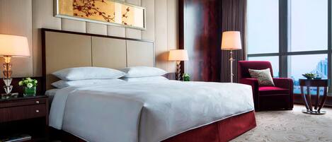 A hotel room with a large bed, two bedside lamps, a red armchair, and a view of the city through large windows.