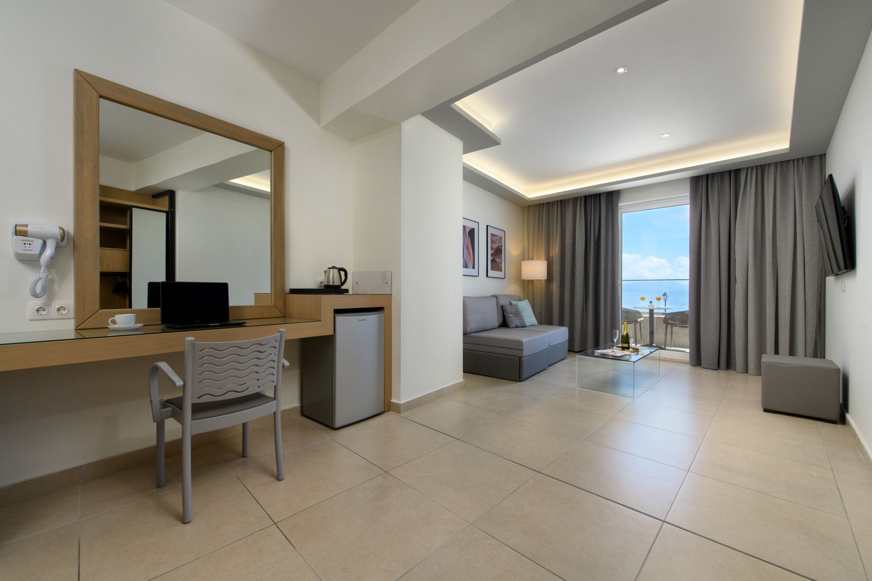 A modern hotel room with a desk, chair, and a small refrigerator. There is a sofa and a television mounted on the wall.