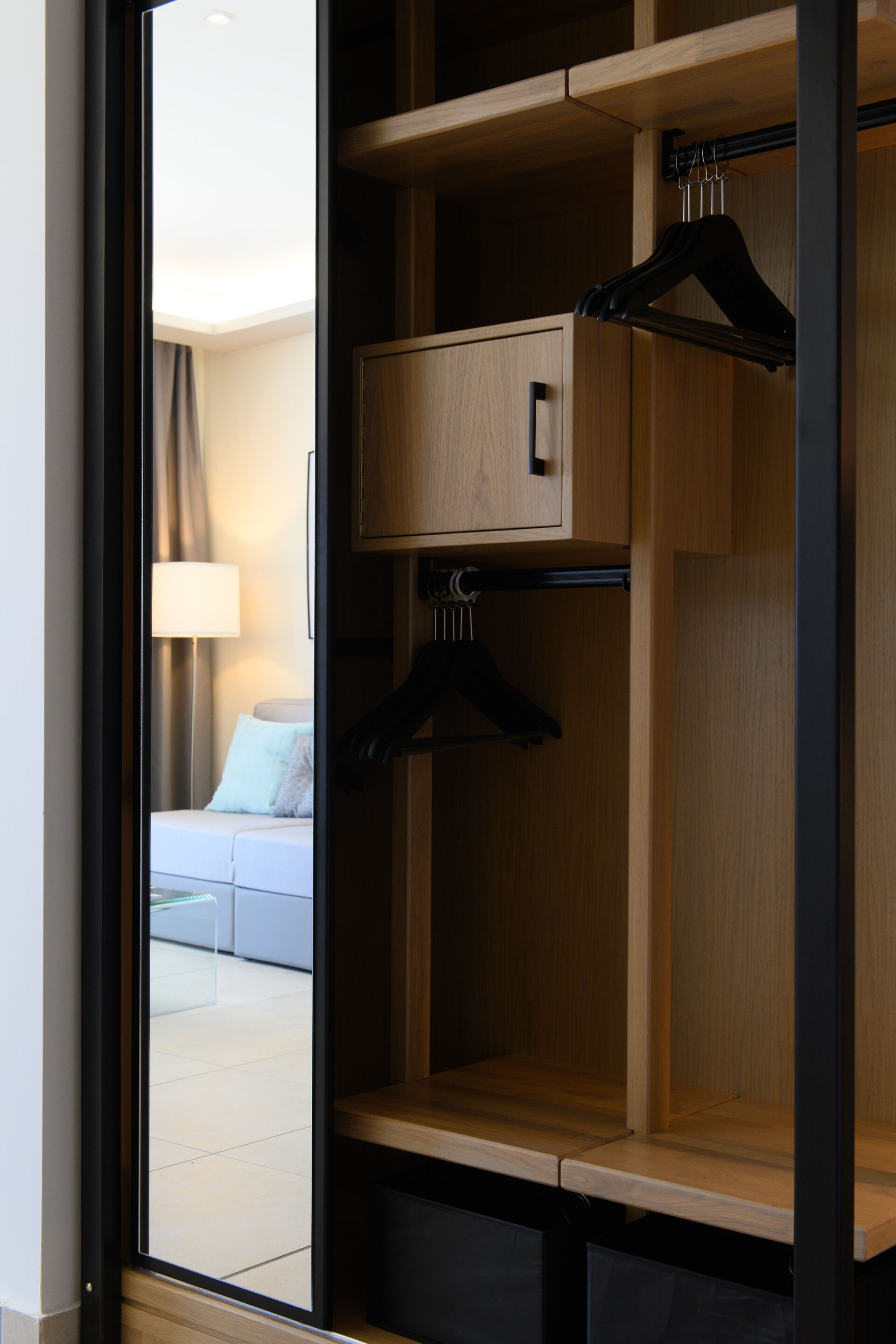 A hotel room with a wooden wardrobe, a hanging closet, and a view of a living area with a sofa and lamp.