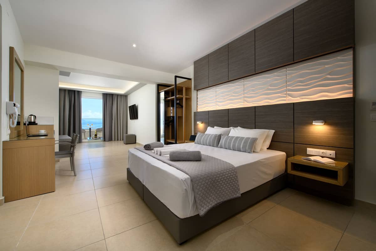 A modern hotel room with a large bed, a desk, and a view of the sea.