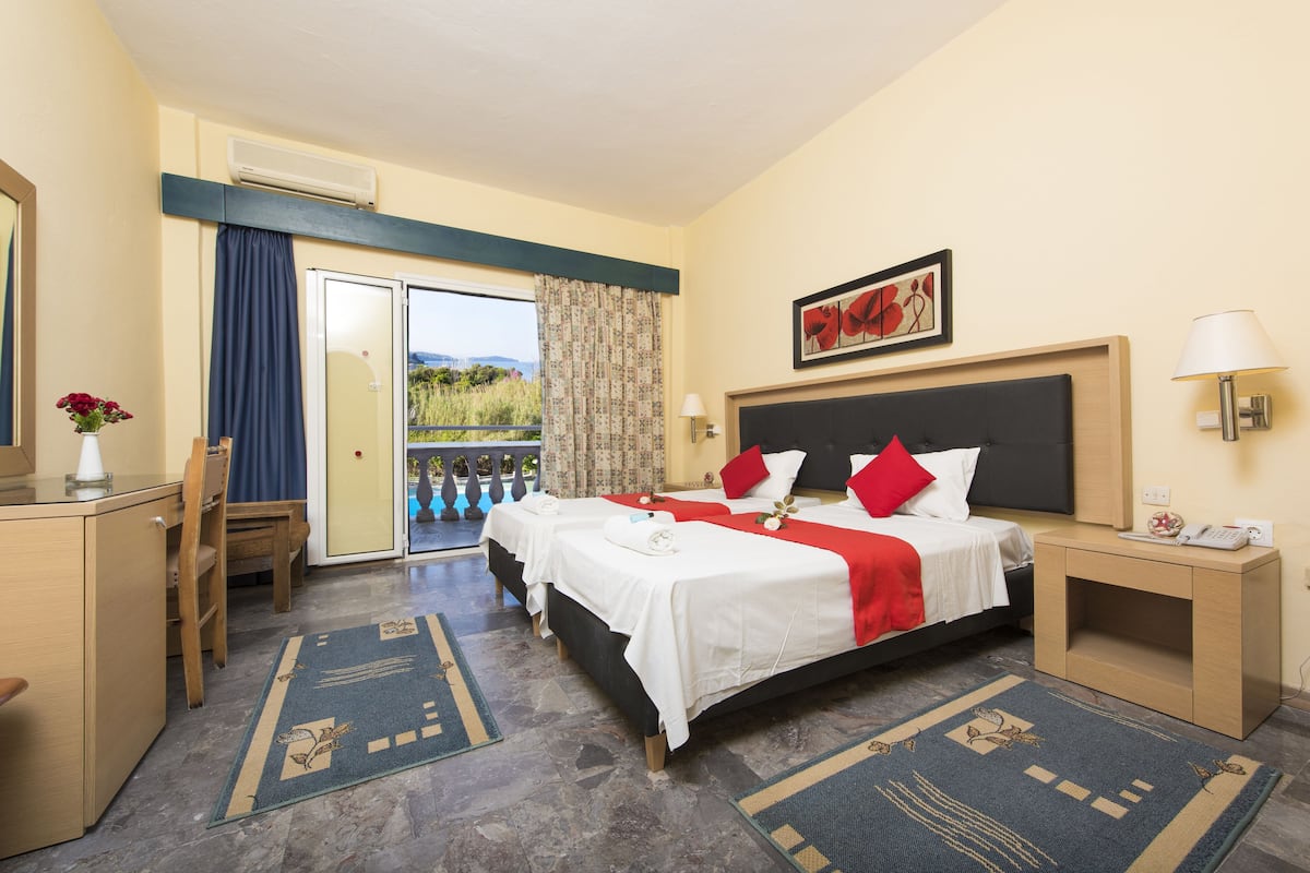 A hotel room with a large bed, two bedside tables, a desk, and a chair. There is a window with curtains, a painting on the wall, and a view of the outdoors.
