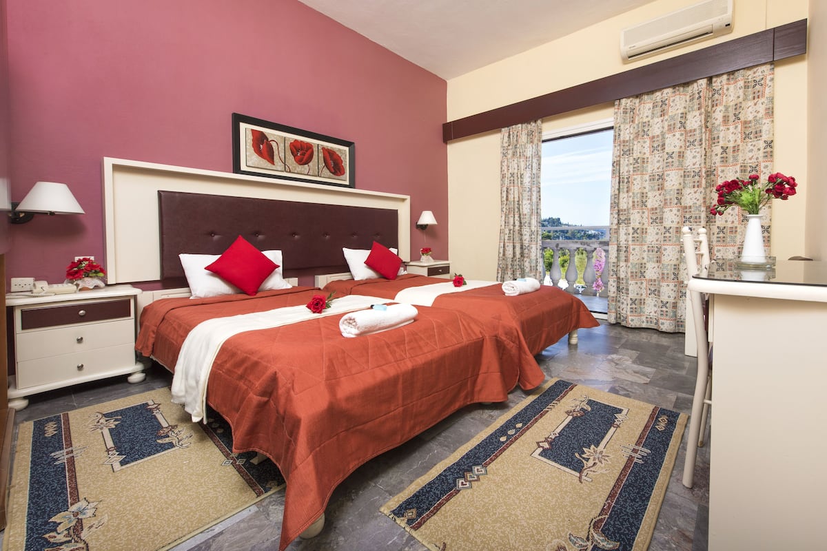 A hotel room with two beds, a desk, a chair, a window with curtains, and a view of the city.