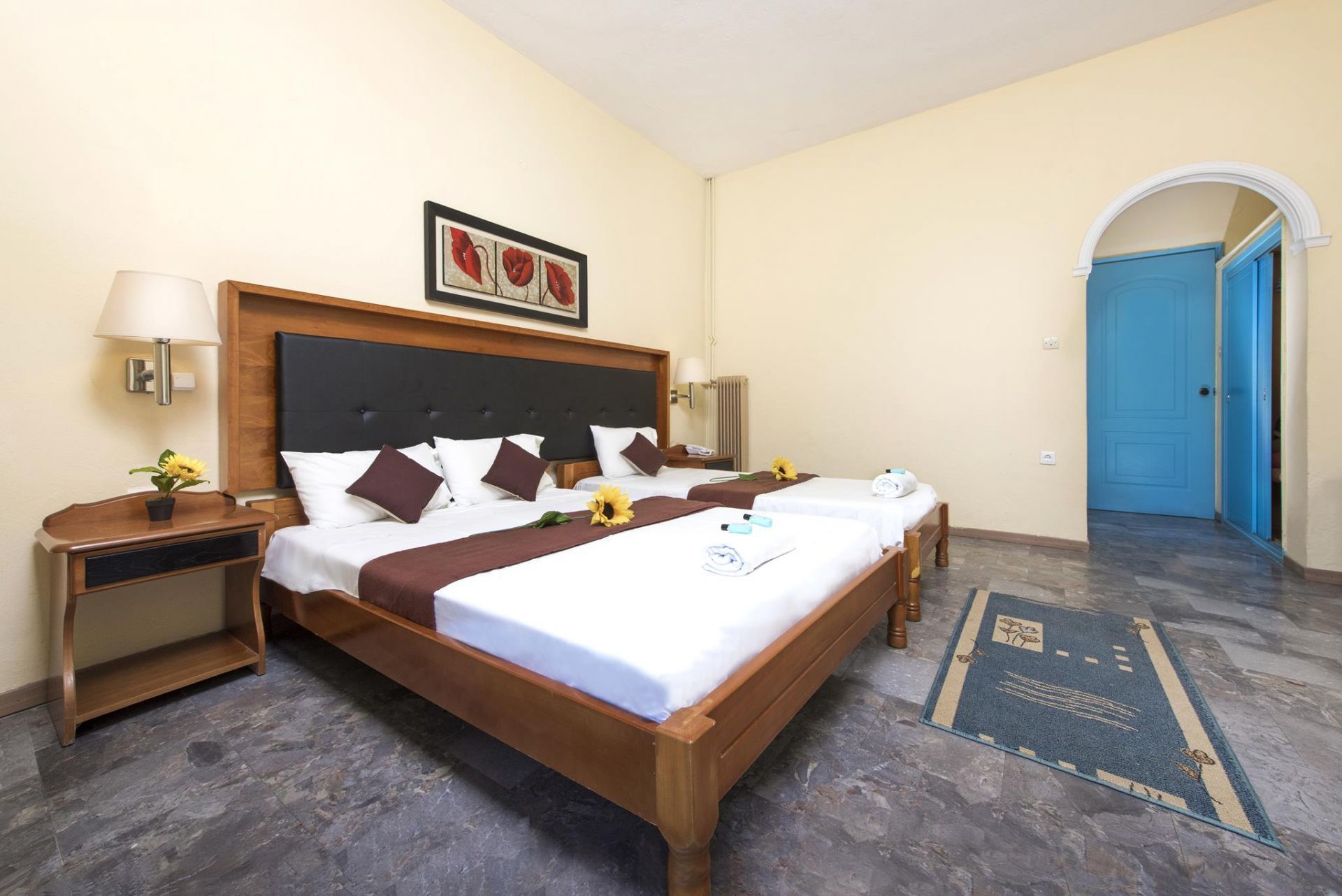 A hotel room with a large bed, two bedside tables, a nightstand, a lamp, a painting, and a blue door.
