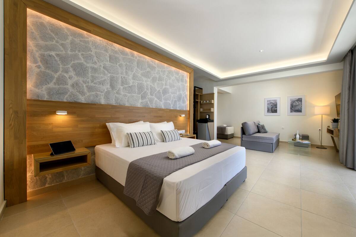 A modern hotel room with a large bed, a wooden headboard, a stone accent wall, a bedside table with a laptop, a gray sofa, and a small table with a lamp.