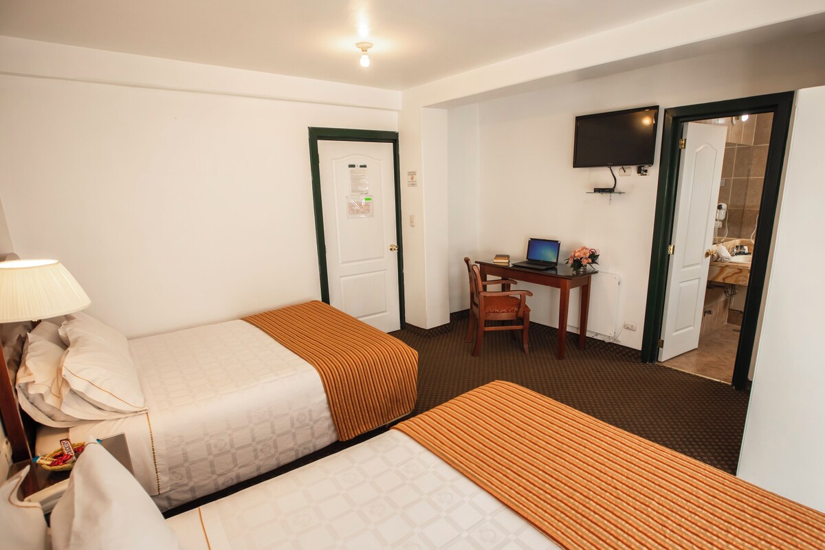 A hotel room with two beds, a desk with a laptop, a TV, and a door leading to a bathroom.