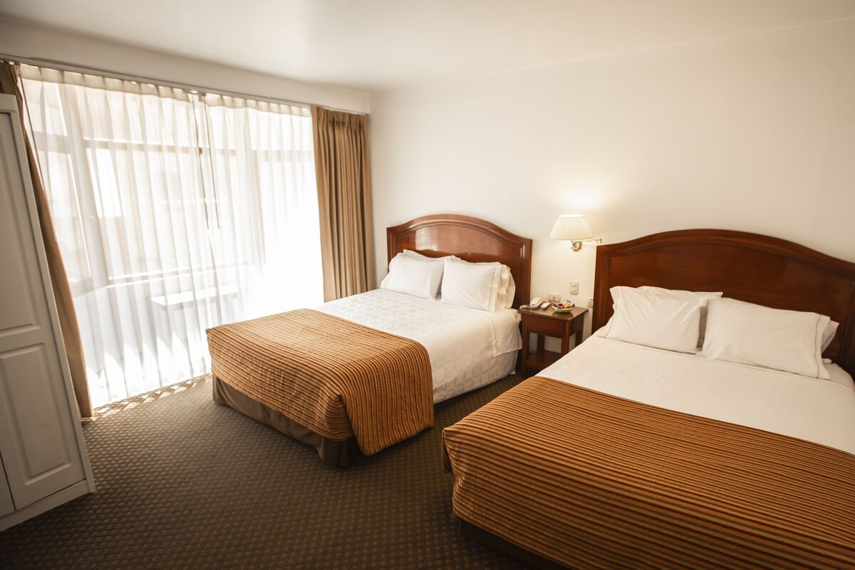 A hotel room with two beds, a window with curtains, a bedside table with a lamp, and a carpeted floor.