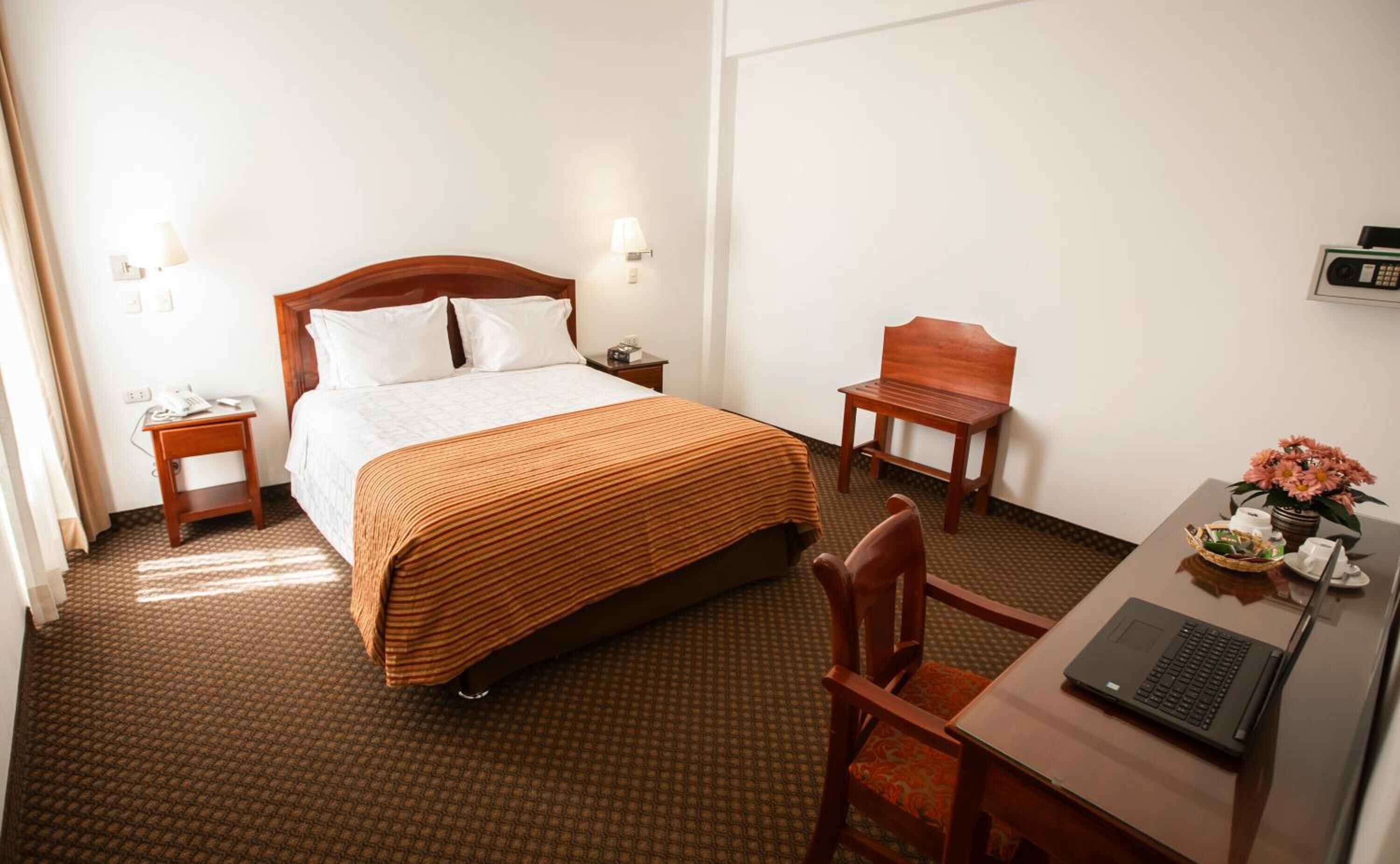 A hotel room with a bed, bedside tables, a desk with a laptop, a chair, and a small table with flowers.