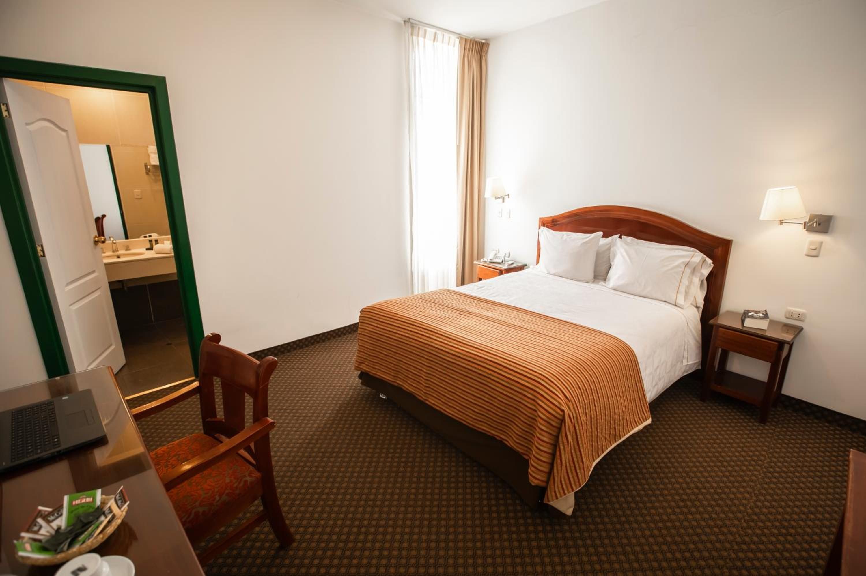 A hotel room with a bed, a desk, a chair, a nightstand, and a bathroom visible through an open door.