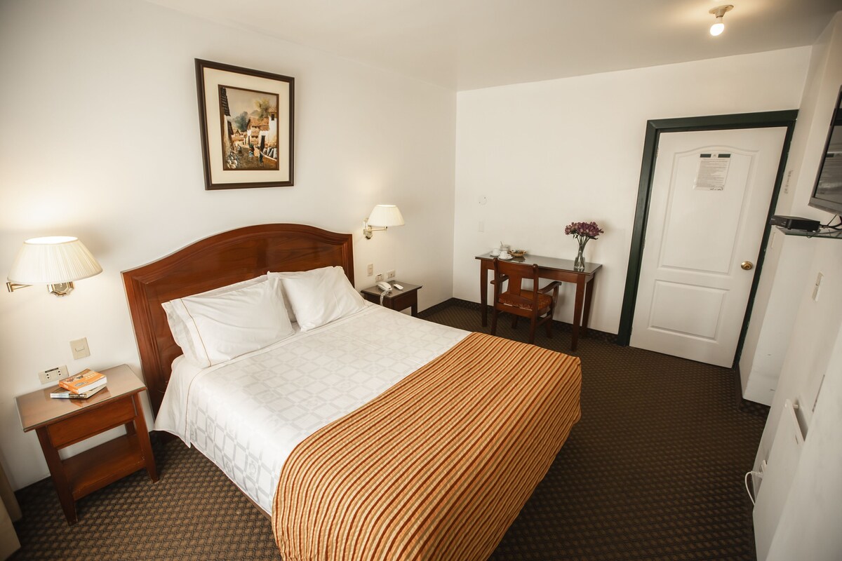 A hotel room with a bed, bedside tables, a desk, a chair, a television, and a door.