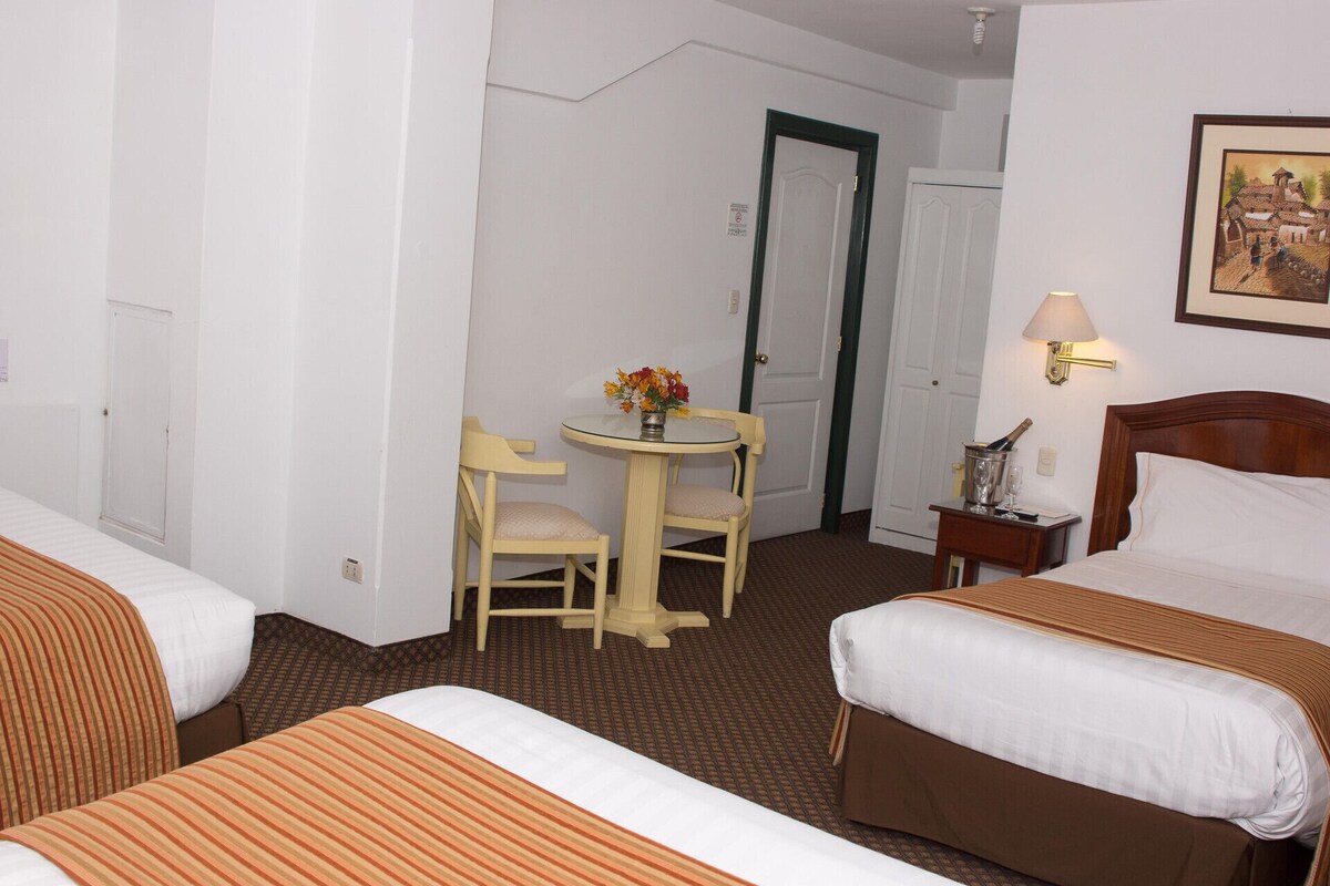 A hotel room with two beds, a small table with a vase of flowers, a chair, a nightstand with a lamp, and a door.