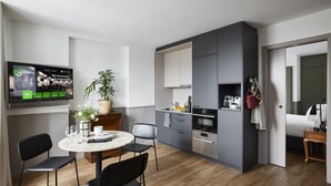A modern kitchen with a built-in oven, a dining area with a round table and chairs, and a TV mounted on the wall displaying a news channel. - Base Lausanne (Lausanne)