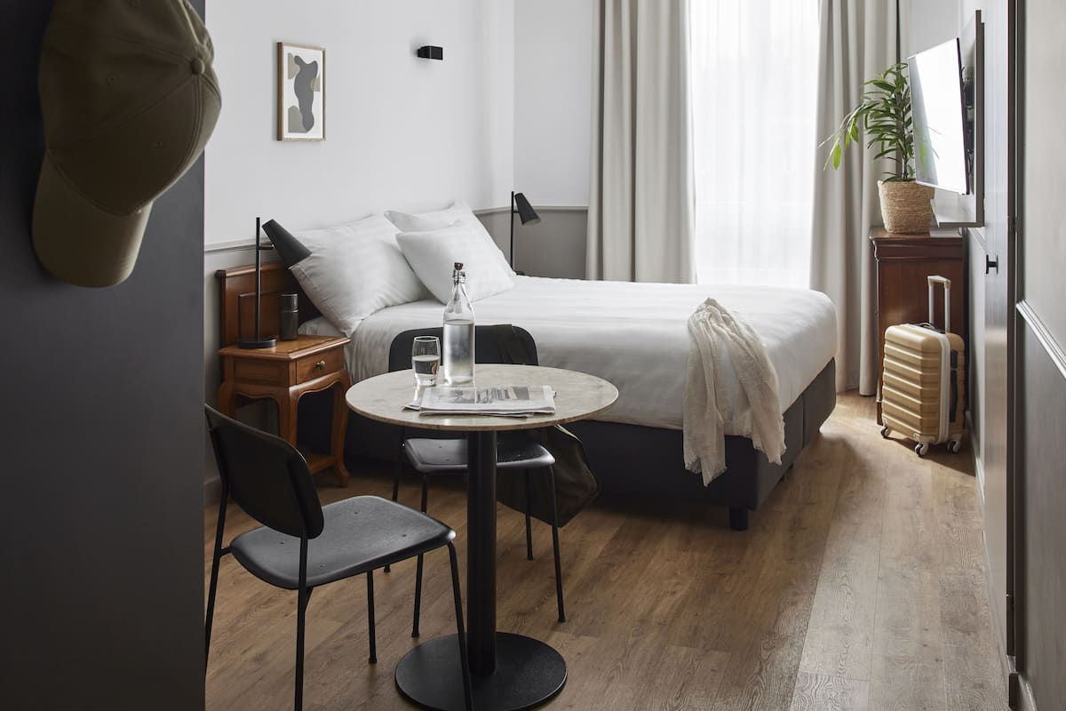 A hotel room with a bed, a round table with chairs, a desk, a suitcase, a potted plant, and a hat on the wall.