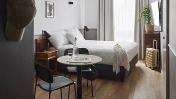 Une chambre d’hôtel avec un lit, une table ronde entourée de chaises, un bureau, une valise, une plante en pot et un chapeau accroché au mur.