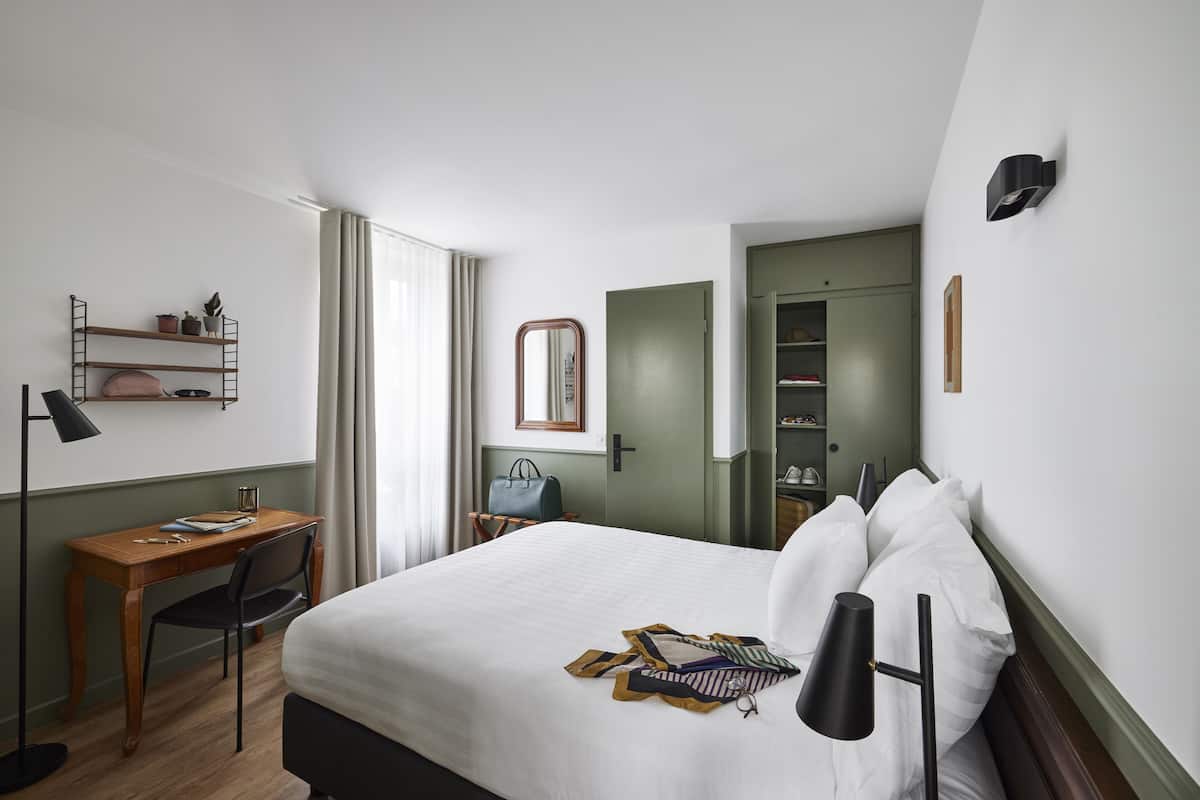 A hotel room with a bed, a desk, a chair, a lamp, a wardrobe, and a mirror.