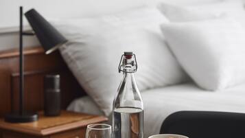 Ein Hotelzimmer mit einem Bett, einem Nachttisch mit Lampe, einer Wasserflasche, einem Glas, einer Schüssel mit Müsli und einer Zeitung auf dem Tisch.