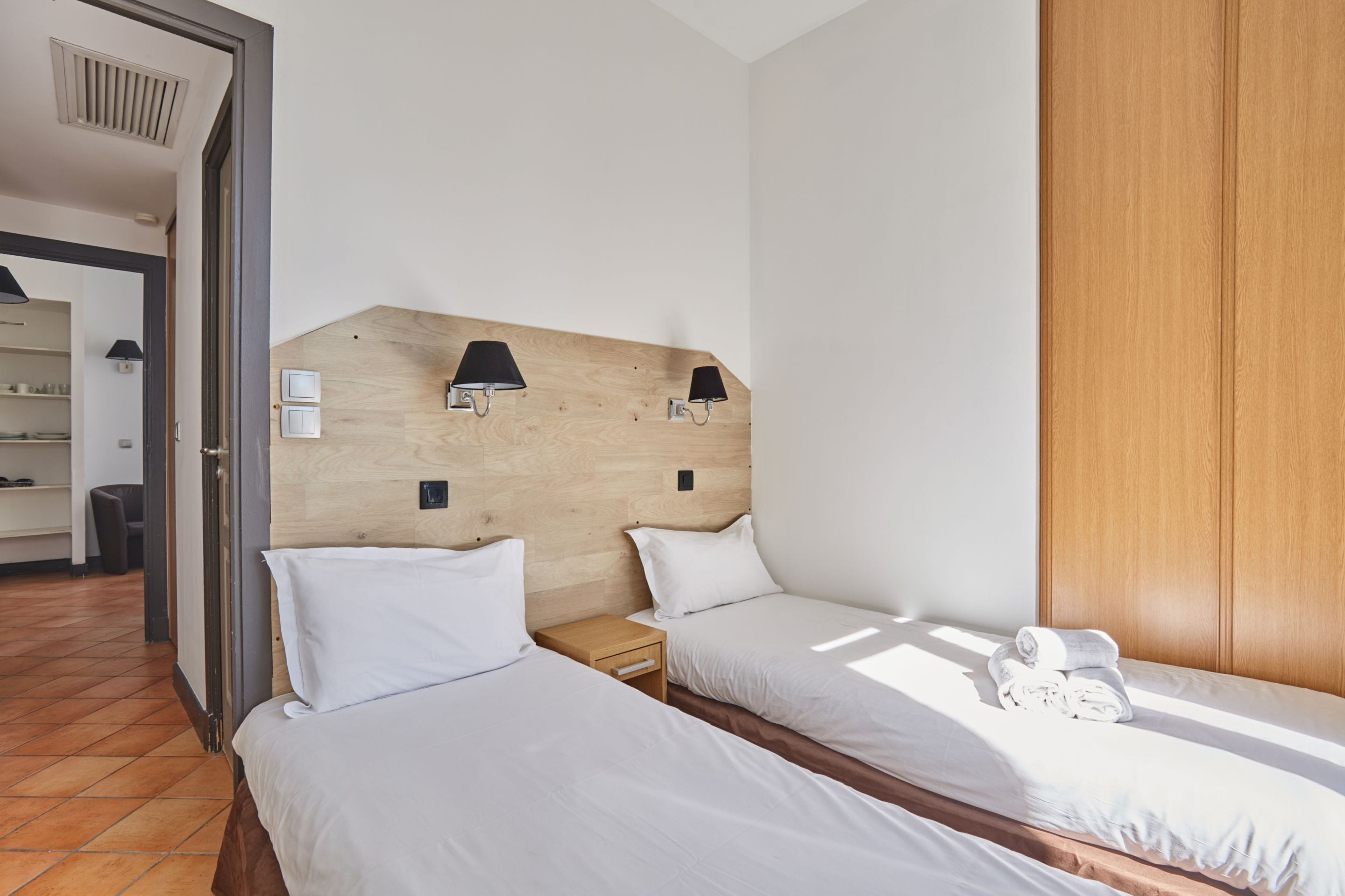 A hotel room with two single beds, a wooden headboard, a wooden wardrobe, and a small wooden nightstand with a lamp.