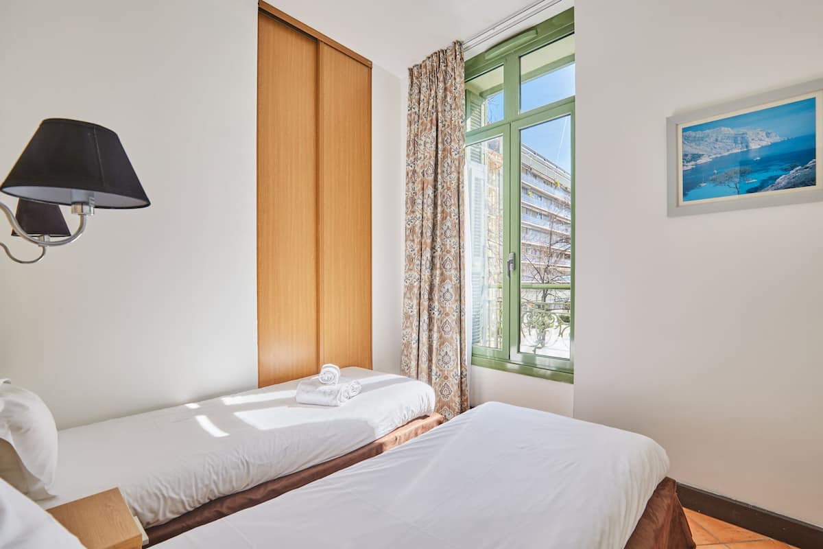 Habitación de hotel con dos camas, una ventana con cortinas, una lámpara de pared y un cuadro enmarcado.