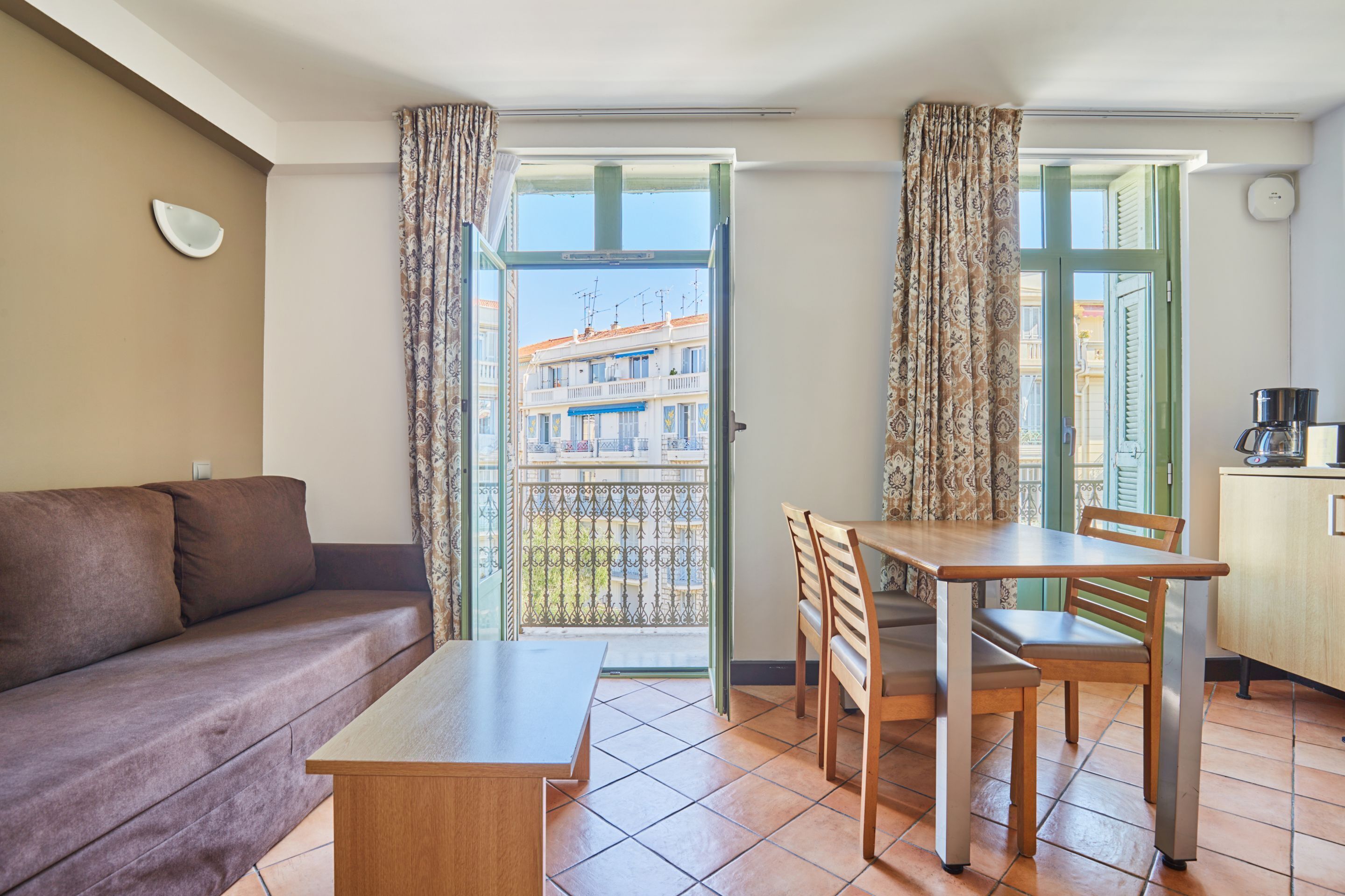A living area with a sofa, a dining table with chairs, a kitchen area, and a balcony with a view of buildings.