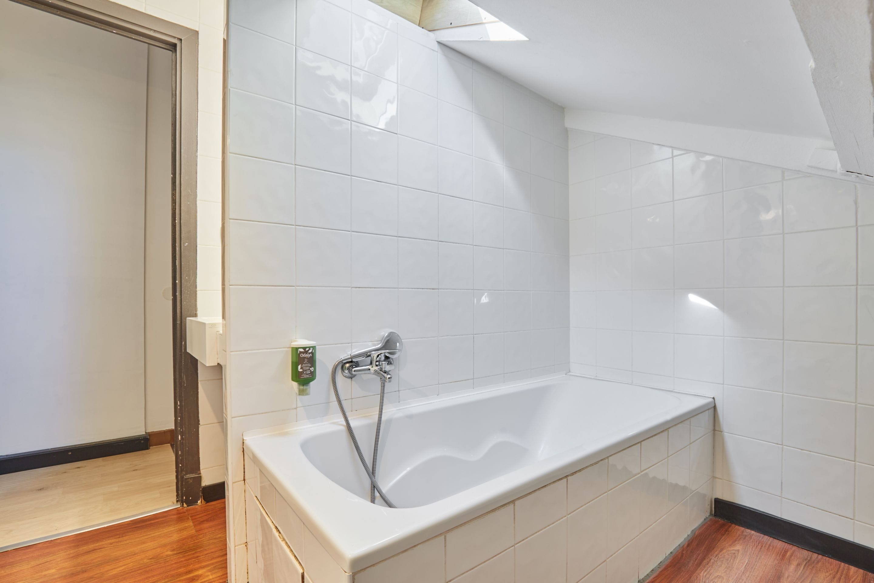 Un baño con bañera, baldosas blancas, suelo de madera y mandos de ducha visibles.