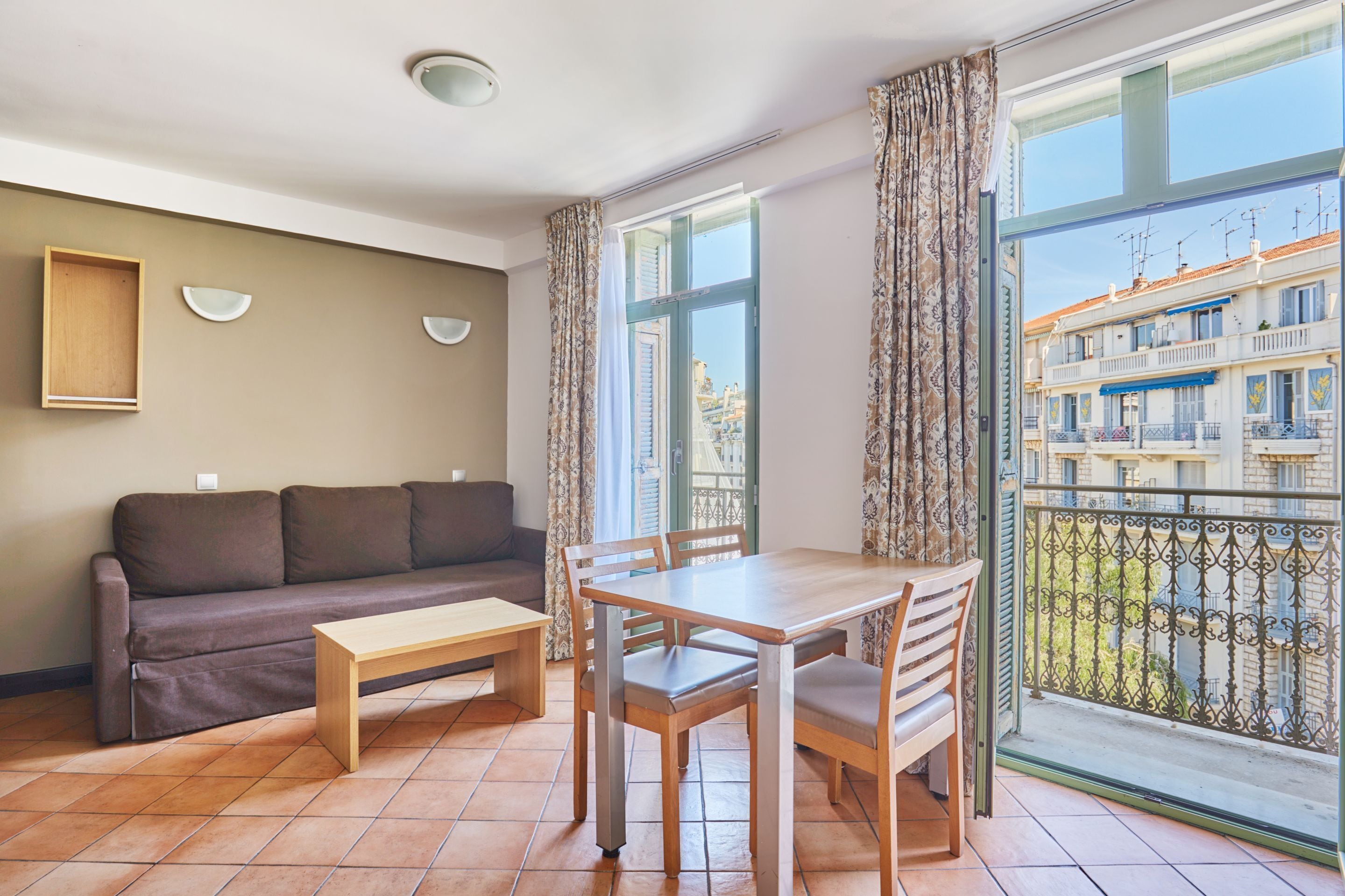 A living room with a sofa, a wooden table, and chairs, a window with curtains, and a view of buildings outside.