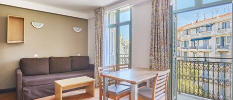 A living room with a sofa, a wooden table, and chairs, a window with curtains, and a view of buildings outside.
