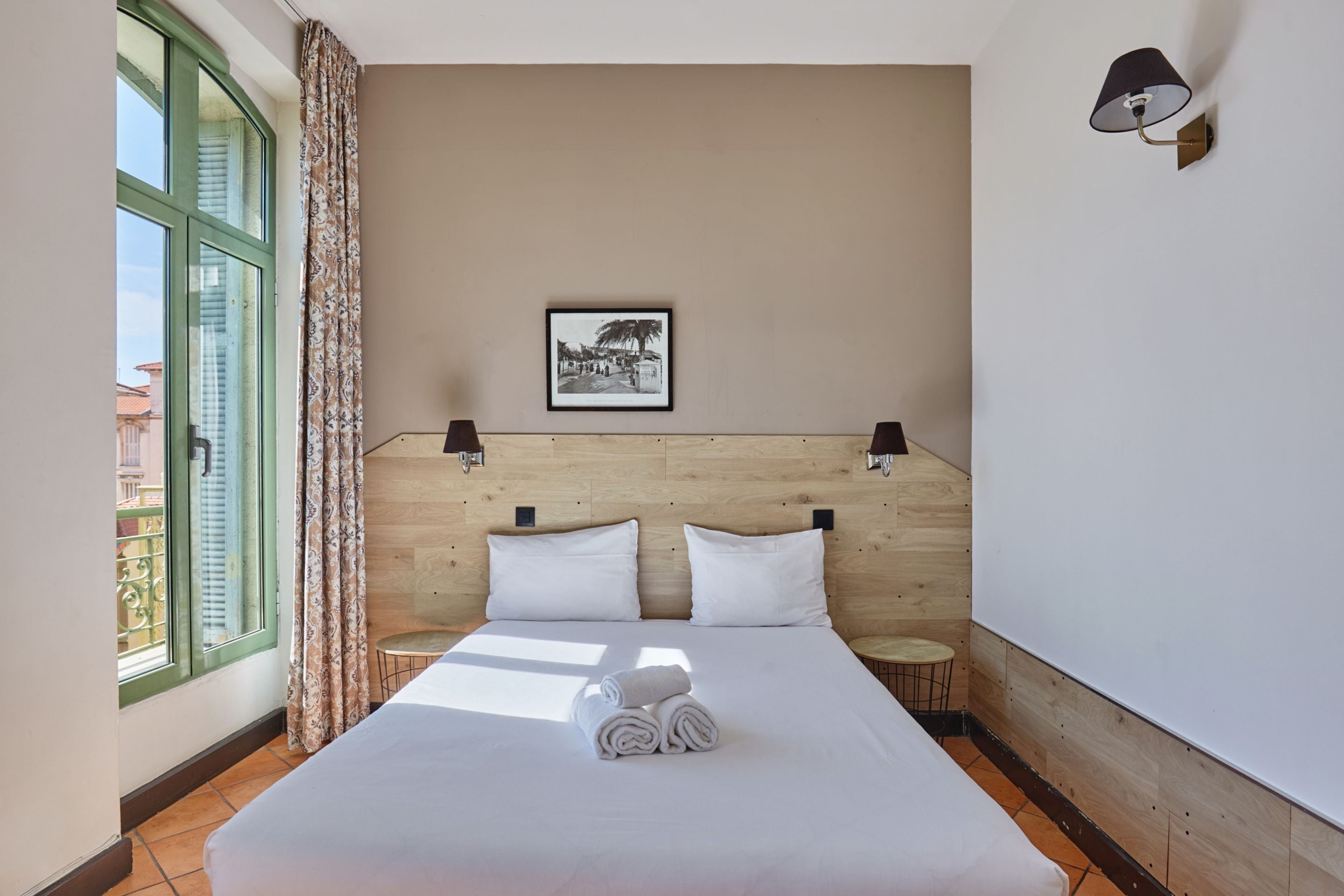 A hotel room with a bed, two bedside tables, a window with curtains, a wall-mounted lamp, and a framed picture on the wall.