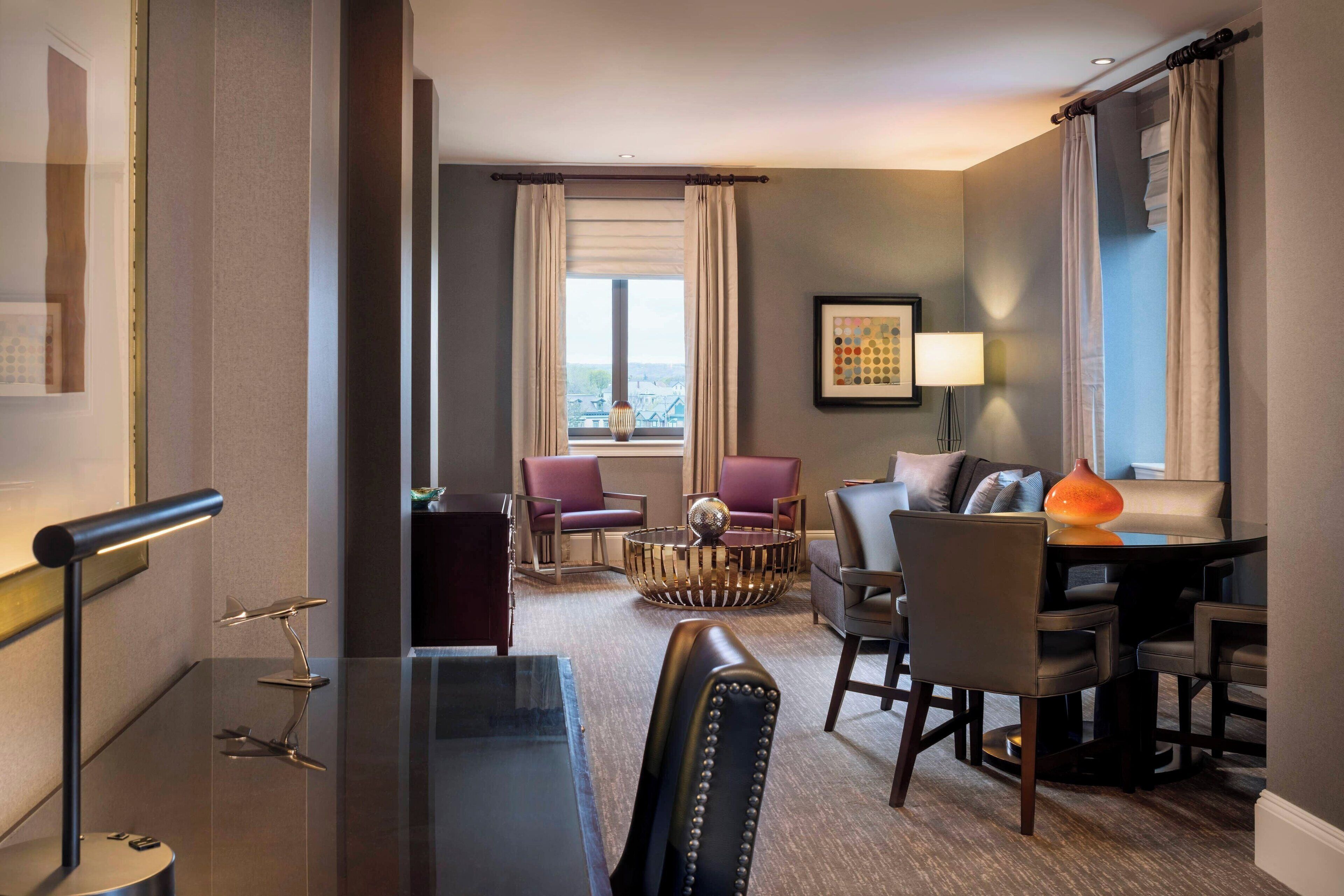 A modern hotel room with a dining area, a sofa set, a round coffee table, and a view of the city through the window.