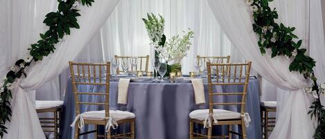 Una mesa dispuesta para una recepción de boda, con cortinas blancas y adornos florales.