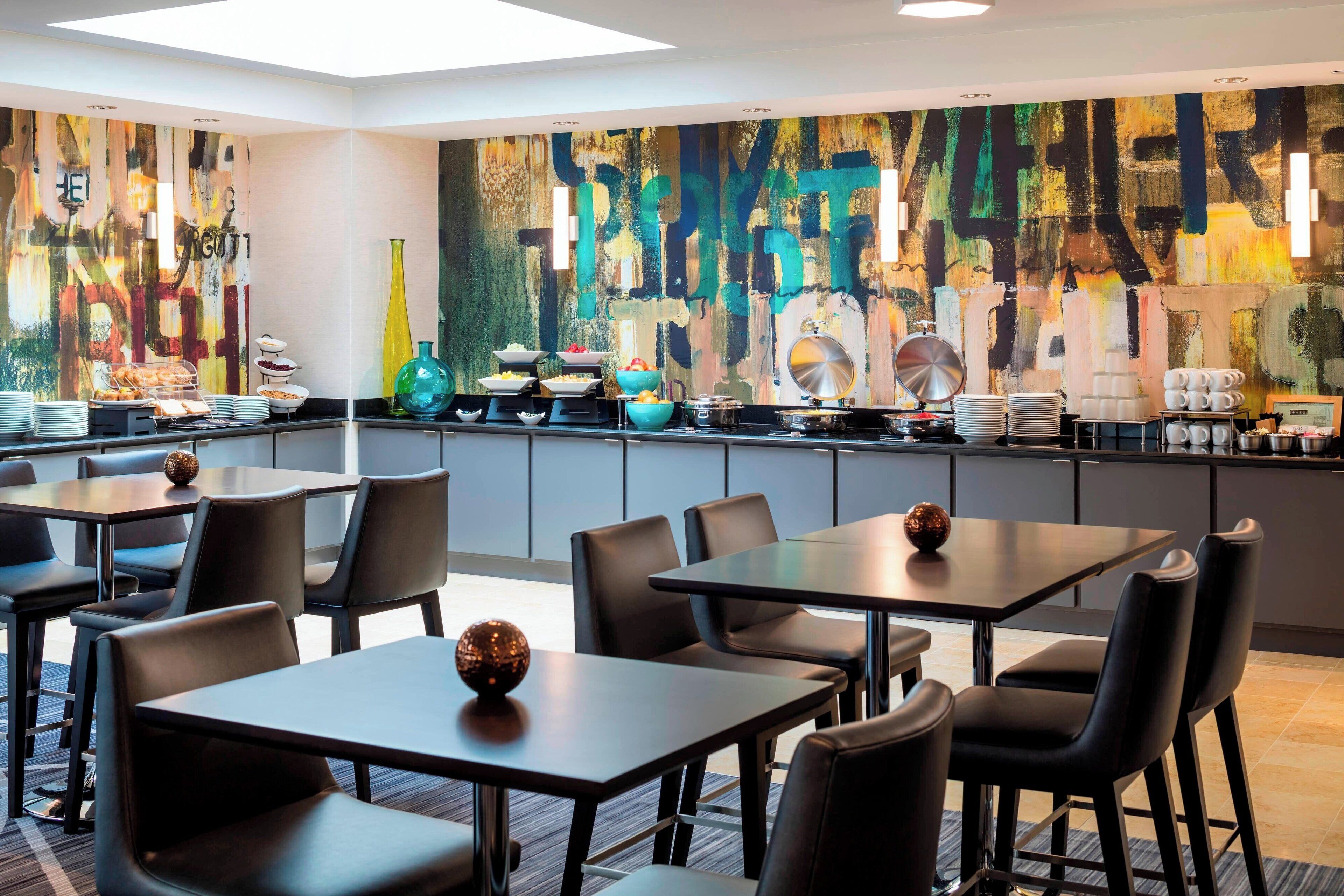 A modern dining area with tables and chairs, a buffet with various dishes, and a colorful abstract mural on the wall.