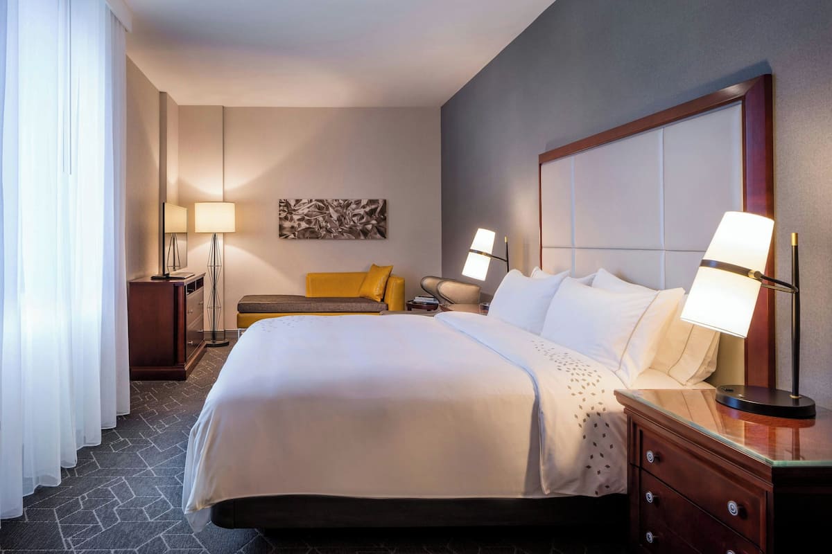 A hotel room with a large bed, two bedside tables with lamps, a nightstand, a sofa, and a wall-mounted artwork.