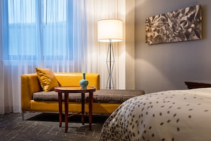 A hotel room with a yellow sofa, a small round table with a vase, a floor lamp, and a bed with a patterned bedspread.