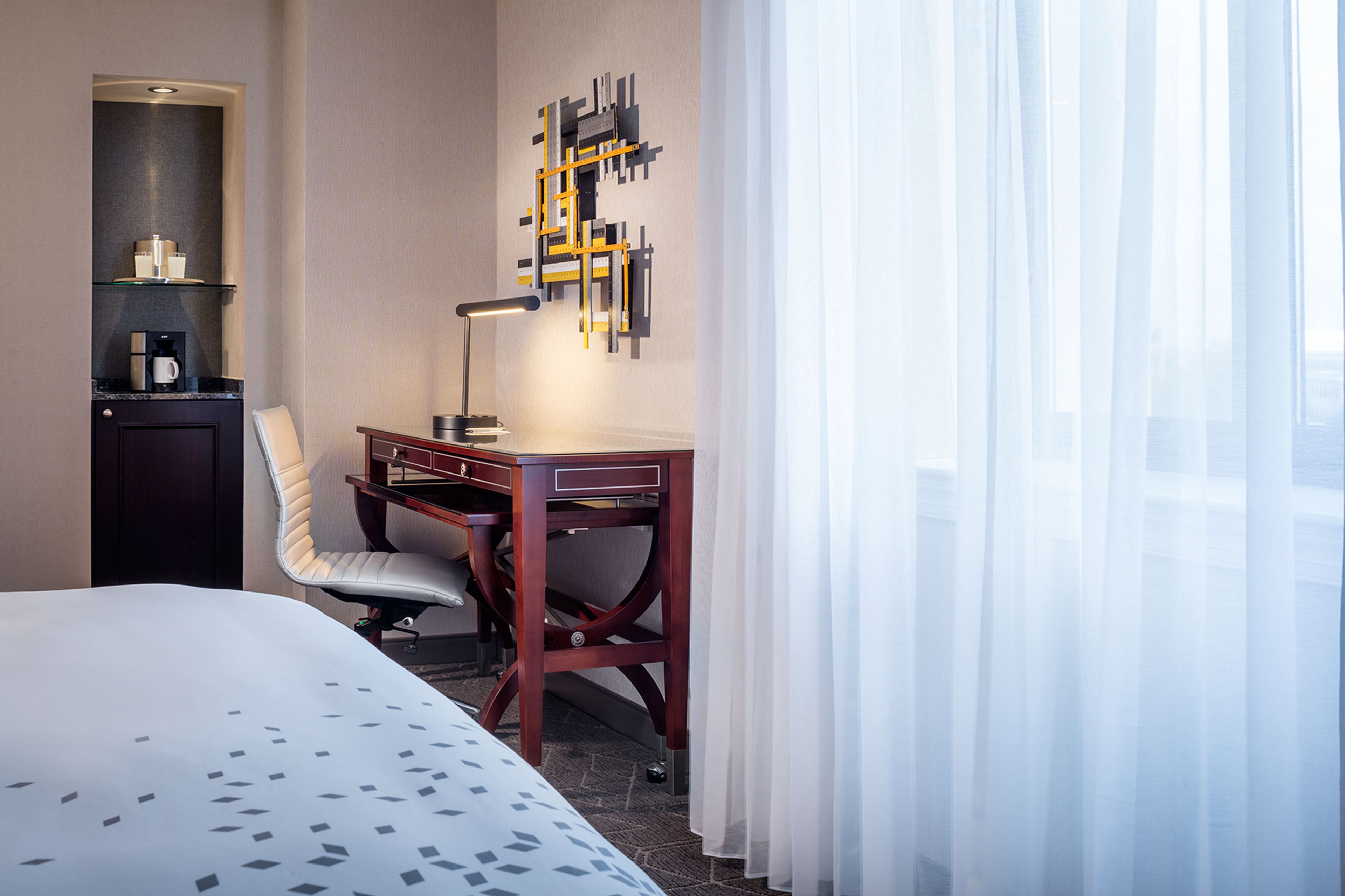 A hotel room with a bed, a desk with a lamp, a chair, a shelf with a coffee maker, and a wall art piece.